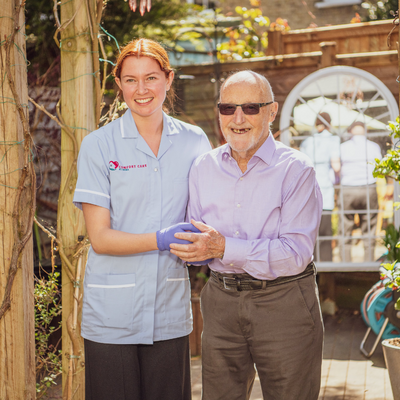 Female care professional assisting older gentleman with motor neurone disease and holding his hand to
help him walk outside